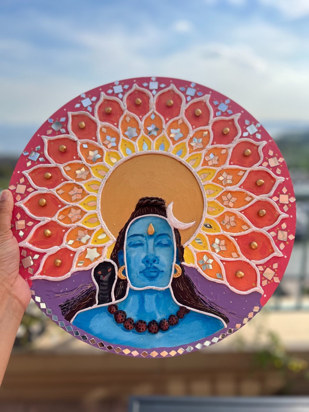 Round Lippan artwork of a blue Shiva with a golden sun behind him, framed by orange petals and mirrors